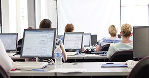 multiple monitor screens sitting on long desks with students at desks facing classroom whiteboard